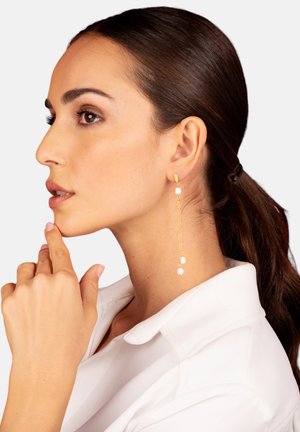 Boucles d'oreilles pendantes dorées dotées de deux petites accents carrés, suspendues à une chaîne délicate, présentées portées par un modèle arborant une queue de cheval lisse.