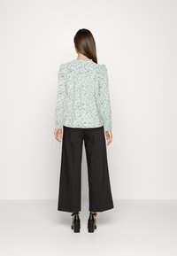 Blusa floreale verde chiaro con maniche lunghe, dettagli arricciati sulle spalle, abbinata a pantaloni neri a gamba larga e tacchi neri con cinturino alla caviglia.