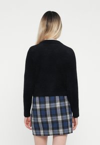 Schwarze, flauschige, kurz geschnittene Strickjacke mit langen Ärmeln, kombiniert mit einem karierten Rock in Blau, Schwarz und Weiß und mit strukturiertem Finish.