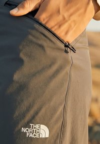 Ruka upravujúca zips na sivých outdoorových nohaviciach The North Face, zobrazených v páse s viditeľným logom a detailným záberom na textúru látky.