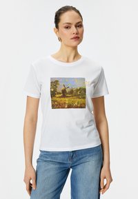 Camiseta blanca de algodón con un estampado de paisaje colorido que presenta un molino de viento, enmarcado por árboles verdes y un cielo amarillo, acentuado por la palabra "Artista."
