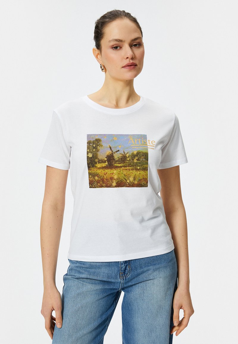 Hvid bomulds t-shirt med et farverigt landskabsprint, der viser en vindmølle, indrammet af grønne træer og en gul himmel, akkompagneret af ordet "Artiste."
