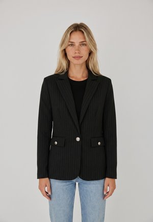 Donna bionda che indossa un blazer nero a righe sottili con bottoni argento sopra una maglietta nera e jeans azzurri, in piedi davanti a uno sfondo semplice.