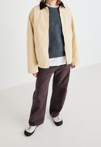 Beżowa kurtka na zamek z czarnym kołnierzem, pod spodem szary prążkowany sweter, brązowe spodnie o szerokich nogawkach oraz czarno-białe sportowe buty.