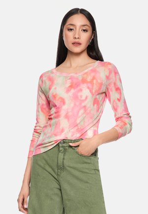 Frau mit langem dunklem Haar, die ein langärmeliges Oberteil mit rosa-weißem Muster und hoch taillierte olivgrüne Hose trägt, mit einer Hand in der Hosentasche.