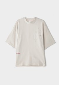 Ljusgrå oversized T-shirt med korta ärmar, med ett litet svart tryck på bröstet och en röd logotyp vid fållen. Mjuk bomullsmaterial.