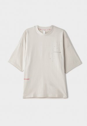 Hellgrau oversized T-Shirt mit kurzen Ärmeln, mit einem kleinen schwarzen Druck auf der Brust und einem roten Logo am Saum. Weiches Baumwollmaterial.