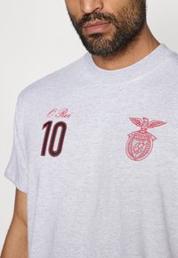 Γκρίζα βαμβακερή μπλούζα T-shirt �με κόκκινο κομμένο γραφικό που περιλαμβάνει λογότυπο και "10" στην αριστερή πλευρά του στήθους. Στρογγυλή λαιμόκοψη και κοντά μανίκια.