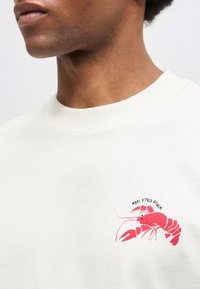 Κοντινό πλάνο ατόμου που φοράει λευκό πουκάμισο με κόκκινο γραφικό αστακού και κείμενο "Marc O'Polo Denim" στο στήθος.