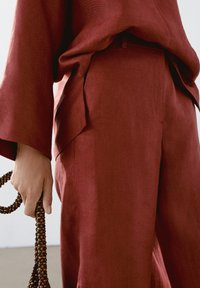 Pantalon en lin rouge à coupe décontractée, avec un pli latéral et un devant plat. Une main tient une sangle accessoire ornée de perles.