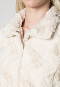 Chaqueta de piel sintética color crema con una superficie texturizada; presenta un amplio cuello y un brillo sutil. Vista en primer plano de la caída del tejido.