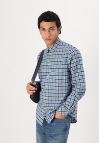 Checkered blue and white button-up shirt with a collar, long sleeves, and a chest pocket, paired with denim jeans and a black shoulder bag.