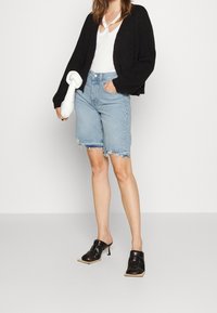 Cardigan en maille noire, haut blanc avec détail découpé, shorts en denim bleu clair avec ourlet effiloché et mules à talons noirs.