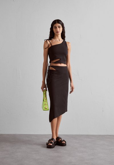 Femme portant un haut noir asymétrique et une jupe avec des découpes, tenant un sac à main vert avec des accents dorés, se tenant devant un mur gris.