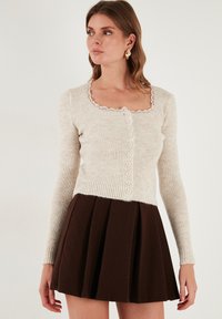Šviesiai béžinė raukšlėta kardiganas su ilgomis rankovėmis, kvadratiniu iškirpimu, nėriniu tinkliniu apdaila ir mygtukų uždarymu, derinamas su rudomis klostuotomis sijonais.