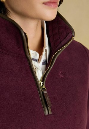 Burgunderroter Fleece-Pullover mit einem gesteppten Kragen, Reißverschluss vorne und braunen Akzenten. Verfügt über goldene Hardware und ein kleines Logo auf der Brust.