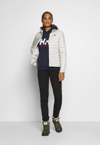 Srebrna pikowana kurtka z kołnierzem stojącym, noszona na granatowym hoodie z czerwonym logo, zestawiona z czarnymi spodniami i ciemnozielonymi butami.