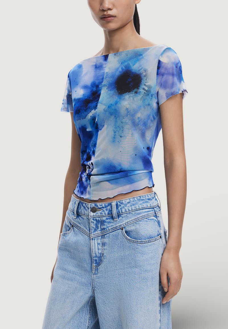 Blå abstrakt mönster kortärmad topp med en något vågig fåll; tillverkad av lätt, slät tyg. Kombinerad med högmidjade jeans.