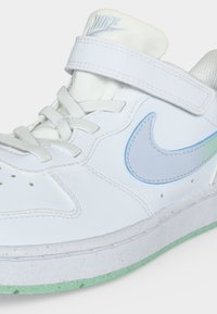 Λευκό υψηλό αθλητικό παπούτσι Nike με ανοιχτό μπλε σήμα, λουράκι με βέλκρο, λευκά κορδόνια και πράσινη απόχρωση στη λεπτομέρεια της σόλας, εμφανιζόμενο από πλάγια μπροστινή γωνία.