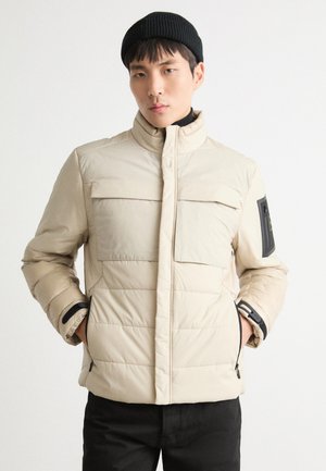 Beige puffjacka med hög krage, horisontell söm, två framfickor och en svart accentpatch på ärmen. Svart stickad mössa.