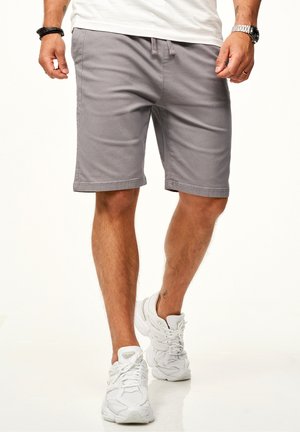 Mann trägt graue Shorts, weiße Turnschuhe, ein weißes T-Shirt, eine Armbanduhr, einen Ring und schwarze Armbänder und steht mit einem Fuß nach vorne.