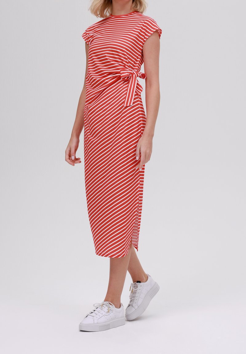 Femme portant une robe midi rayée rouge et blanche avec un détail de nœud sur le côté et des baskets blanches, debout devant un fond uni clair.