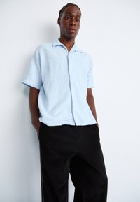 Light blue short-sleeved button-up shirt with a textured fabric, paired with black corduroy pants. Simple design with a collar and no visible patterns.