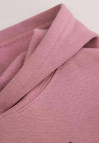 Różowa bawełniana bluza z kapturem o miękkiej fakturze, z złożonym kapturem i prostym wzornictwem z minimalnymi detalami. Materiał wydaje się ciepły i wygodny.