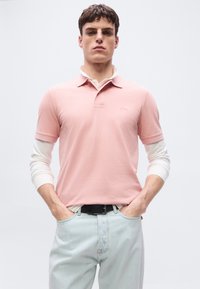 Άνδρας που φοράει ανοιχτό ροζ πόλο πάνω από λευκά μακρυμάνικα και ανοιχτό μπλε τζιν, στέκεται με τα χέρια στις τσέπες σε απλό φόντο.