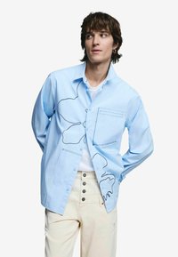 Hele sinine nööbiga särgi, millel on lilleline kontuurimuster, rinnatasku ja pehme puuvillane tekstuur, kantud valge T-särgi peal.