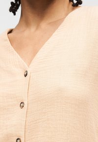 Gros plan d'une personne portant un haut beige texturé boutonné avec un col en V et trois boutons ronds visibles.
