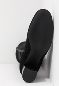 Botte en daim noir positionnée semelle vers le haut contre un coin blanc, montrant une semelle lisse et un talon bloc court avec une semelle en caoutchouc texturée.