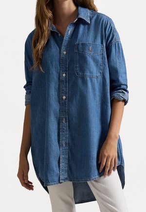 Femme portant une chemise en denim bleu ample à manches longues avec des poignets retroussés et un pantalon blanc, debout devant un fond uni.