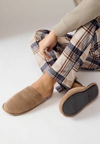 Pantofole slip-on in tessuto intrecciato marrone con parte superiore morbida e suola in gomma, abbinate a pantaloni pigiama a quadri nei toni beige, navy e marrone.