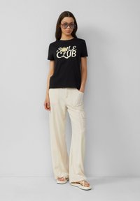 Zwarte katoenen t-shirt met "SMILE CLUB" grafiek in crème, gecombineerd met lichtbeige wijde cargo broek en minimalistische sandalen.