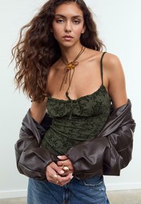 Jeune femme aux longs cheveux bouclés portant un débardeur floral vert, une veste en cuir marron portée sur les épaules, plusieurs anneaux en or et un collier avec un pendentif en forme de fleur.