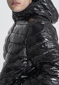 Uomo che indossa un giubbotto imbottito nero lucido con cappuccio e una toppa con logo sul braccio superiore, mostrato dalla vita al mento.