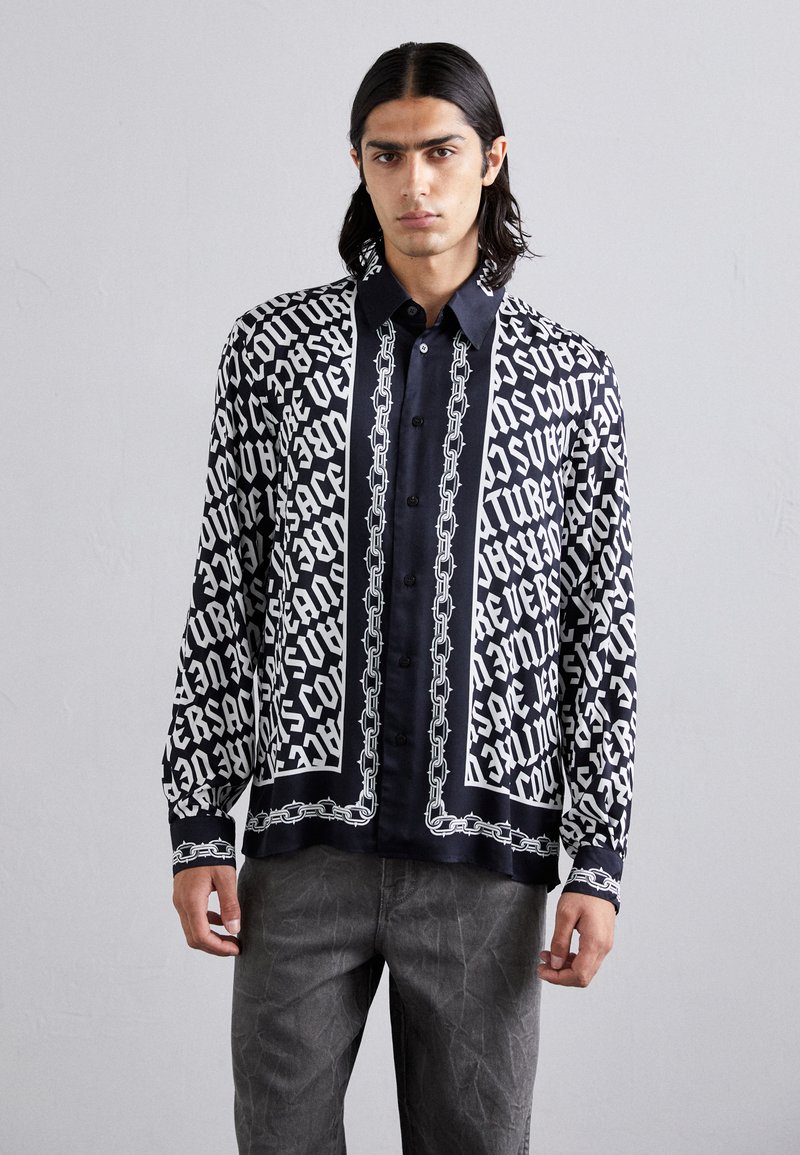 Μαύρο και άσπρο κα patterned μακρυμάνικο πουκάμισο με σχέδιο κειμένου και μοτίβο αλυσίδας, συνδυασμένο με ένα σκούρο πουκάμισο με κουμπιά από κάτω και γκρι παντελόνι.