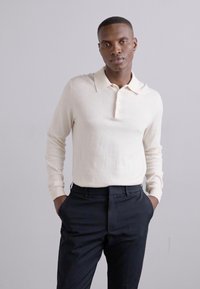 Polo shirt lung cu mâneci lungi, de culoare crem, cu guler și trei butoane, asortat cu pantaloni închiși. Textură netedă, design ajustat.