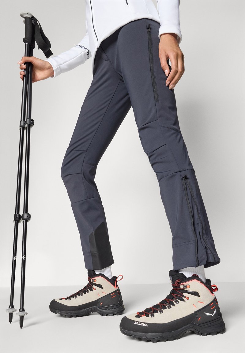 Pantaloni soft-shell grigi con tasche con zip, abbinati a scarponi da trekking beige e neri con accenti rossi e una superficie texture.