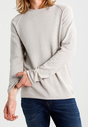 Chemise à manches longues gris clair avec une texture côtelée, des manches raglan et des trous pour les pouces. Associée à un jean bleu. Tissu doux et décontracté.