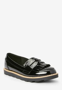 Mocassins en cuir verni noir avec une sangle à franges, bout arrondi et détail latéral texturé, présentant une semelle contrastante avec traction.