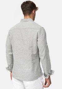 Homme portant une chemise à manches longues grise et blanche à rayures verticales et un pantalon blanc, vu de dos sur un fond uni.