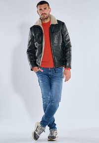 Schwarze Lederjacke mit cremefarbenem Lammfellkragen, rotem geripptem Pullover und blauen, abgenutzten Jeans. Model steht mit den Händen in den Taschen.
