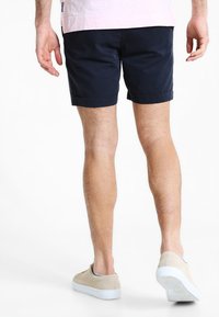 Mørkeblå shorts i bomuld med en foldet kant, iført med lyse beige sneakers mod en ensfarvet hvid baggrund.