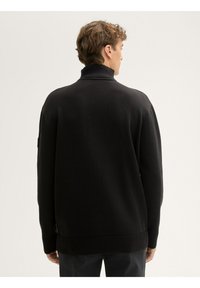 Schwarzer Rollkragenpullover aus strukturiertem Strickstoff. Verfügt über gerippte Bündchen und Saum, mit einem Label an der Seite für zusätzliche Details.