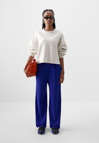 Sweatshirt oversized em creme, calças azuis largas, ténis pretos com acentos roxos e uma mala laranja.