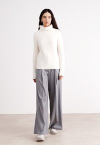 Weißer gerippter Rollkragenpullover kombiniert mit grauen weit geschnittenen Hosen, die eine lockere Passform und eine glatte Textur aufweisen, getragen mit weißen Turnschuhen.