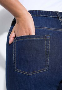 Džínové kalhoty tmavě modré barvy s zadní kapsou zdobenou žlutým stehem. Ruka částečně zasunutá do kapsy.