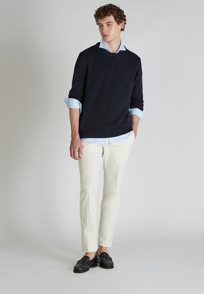 Maglione a maglia blu navy con collo tondo, camicia col cielo chiaro, pantaloni bianchi slim-fit e mocassini neri. Texture liscia e linee pulite.